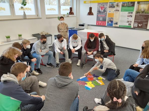 Schülerinnen und Schüler und ihre Lehrerin sitzen im Kreis und schauen, was die französische Lehrerin in der Mitte des Kreises vorstellt